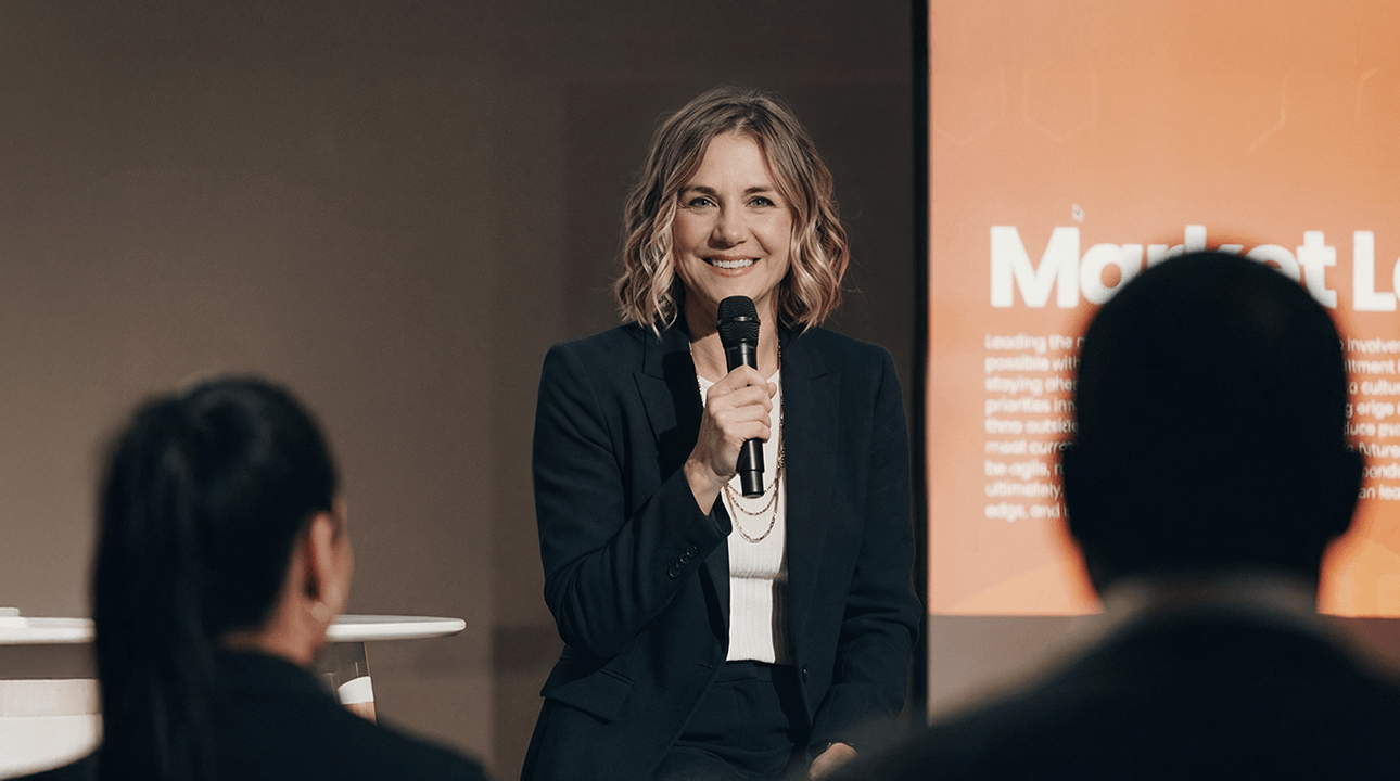 A woman stands confidently holding a tablet, leading a discussion. Audience members listen attentively, one with a hand raised, in a classroom setting.