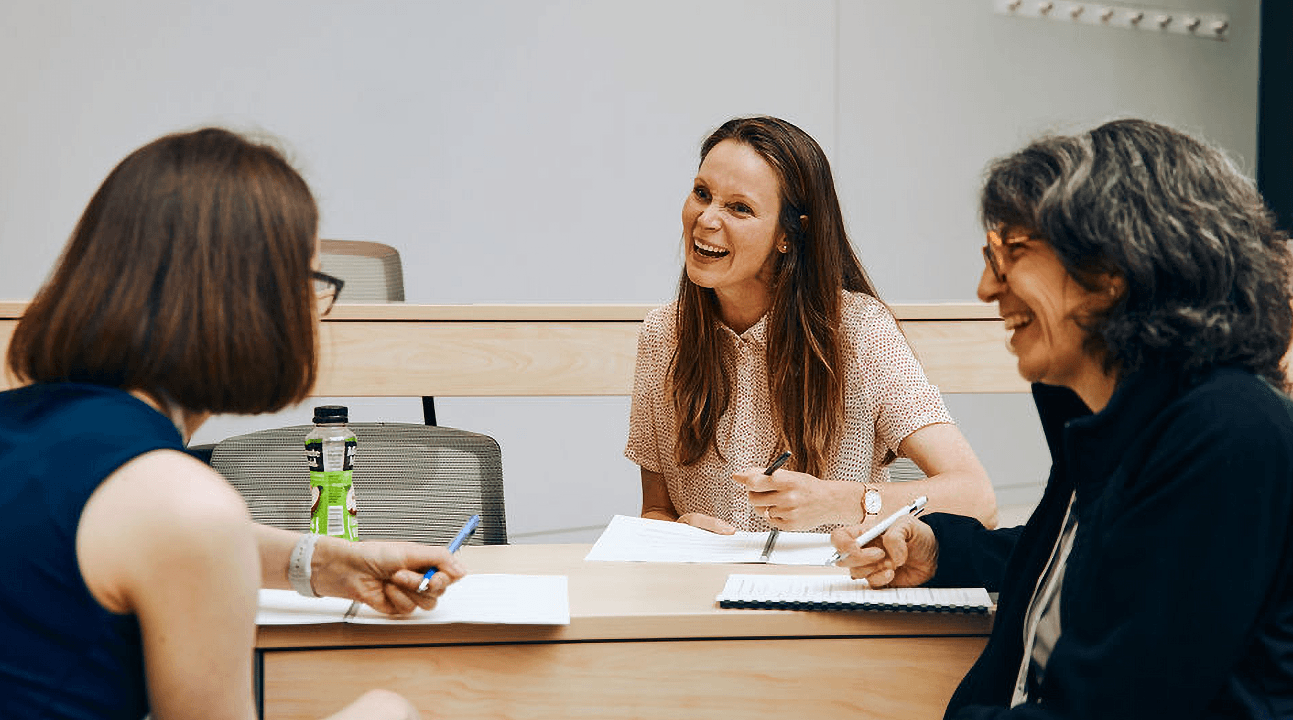 Three women sit at a table in a meeting room, smiling and chatting. They are holding pens and papers, indicating a collaborative discussion.
